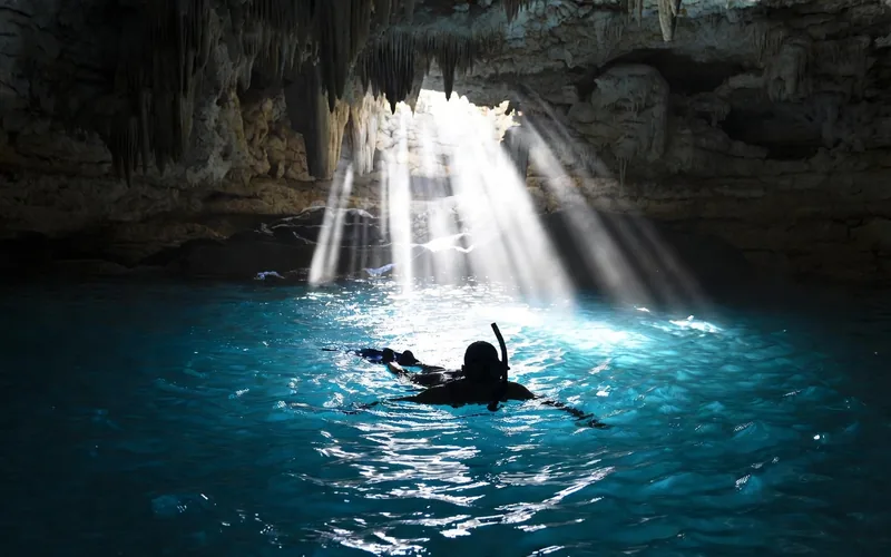 Snorkeler in a cavern cenote with stalactites and light beams near Tulum