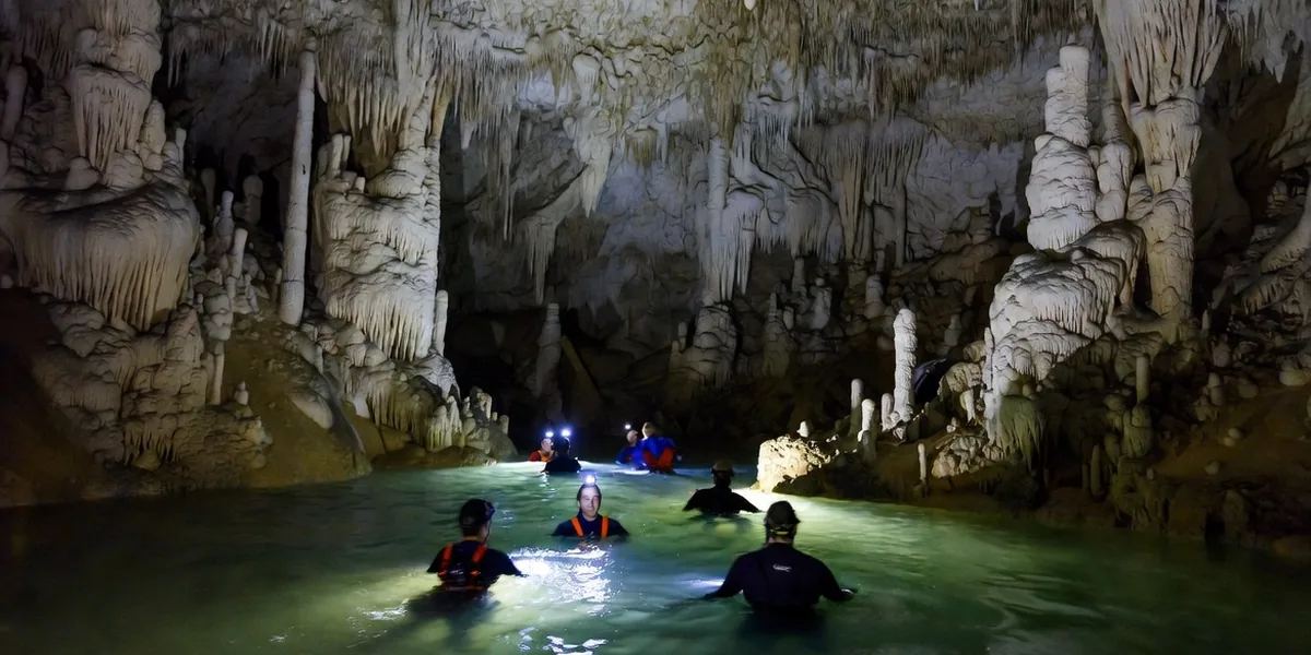 Visitors in helmets and headlamps wading through a stalactite-filled underground river at Río Secreto